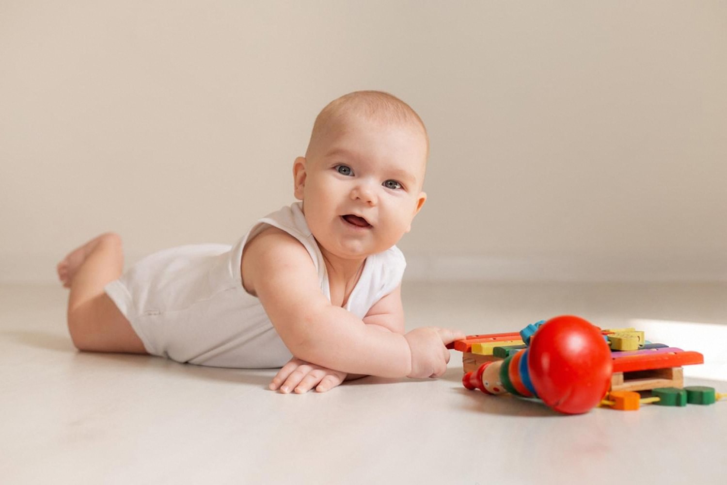 Baby in a white bodysuit lying on the floor beside a wooden xylophone and rattle