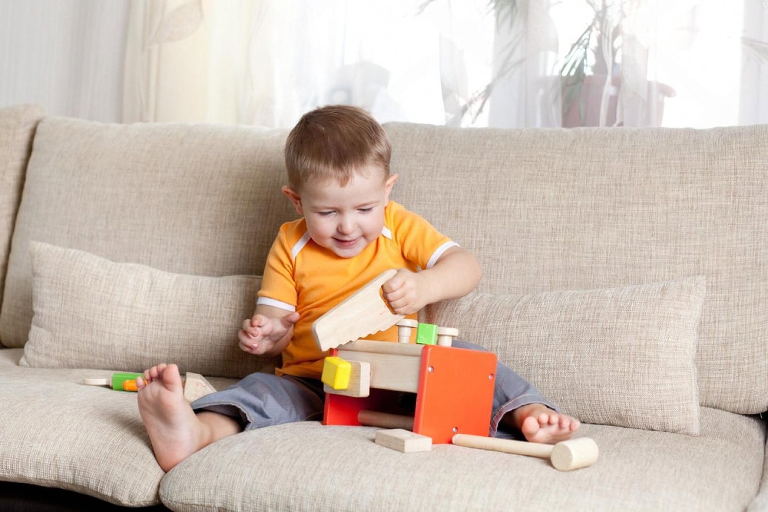 Toddler on a sofa smiling while using a wooden hammer and workbench toy